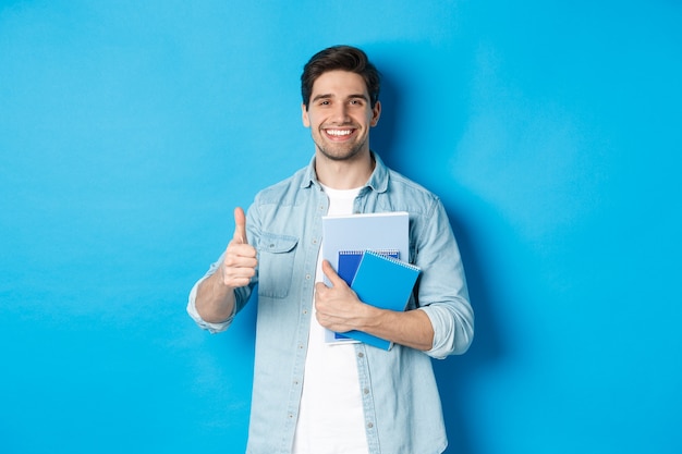 young-man-student-with-notebooks-showing-thumb-up-approval-smiling-satisfied-blue-studio-backg...jpg